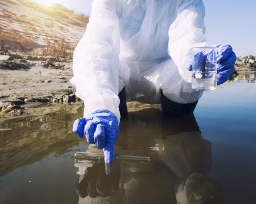 Unrecognizable ecologist taking samples of water with test tube from city river to determine level of contamination and pollution.