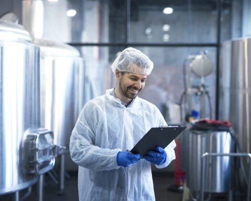 Industrial worker technologist in white suit with hairnet and protective gloves looking at checklist and smiling.