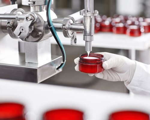 Portrait of big factory process of creating lotion for skincare. Researcher holding container under big metal tube pouring beauty cream isolated over working process at factory and many containers